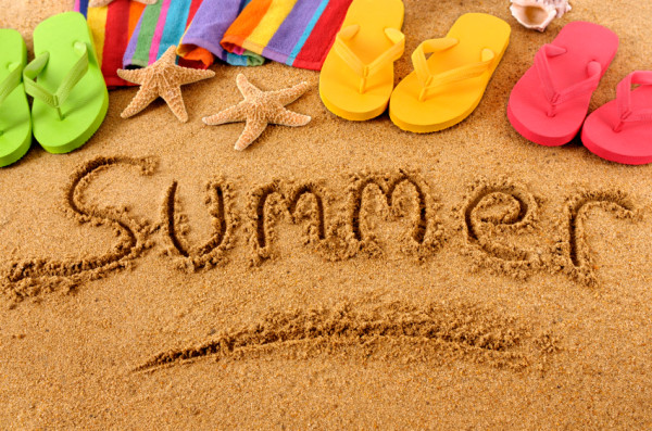 The word summer written in sand and surrounded by flip flops in various colours