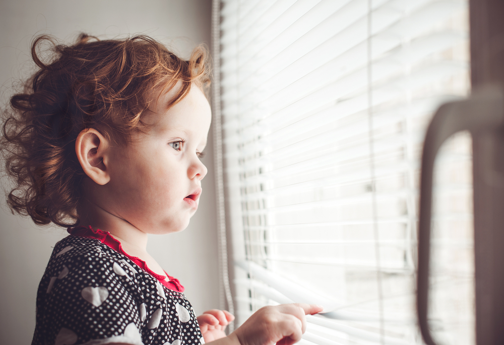 Young toddler with curly red hair looking through blinds on a window